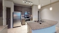 This single-floor residence offers a thoughtfully designed layout with a cohesive blend of modern finishes and functional living spaces. The kitchen serves as a standout feature, equipped with stainless steel appliances including a refrigerator, electric range, and built-in microwave. Dark espresso cabinetry reaches toward the ceiling, complemented by granite countertops, a subway tile backsplash, and large-format tile flooring. A peninsula with an undermount sink and pendant lighting adds both style and practicality, while track lighting illuminates the workspace. Crown molding adds a refined touch throughout. The living area is generously proportioned, featuring a tray ceiling with crown molding detail, a ceiling fan, and plush carpeting. Large windows and glass doors invite abundant natural light and views of the outdoors. The bedroom is carpeted and includes access to a walk-in closet fitted with wire shelving and hanging rods, offering ample storage. The bathroom is well-appointed with a granite-topped dark wood vanity, a large wall mirror, globe vanity lighting, a tiled soaking tub and shower combination, and tile flooring. A double-door pantry or utility closet provides additional storage conveniently located near the kitchen. Neutral tones throughout create a calm, versatile backdrop ready for personalization.