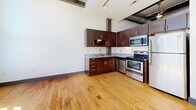This urban loft unit on the third floor at 101 E Sixth St in downtown Winston-Salem showcases an industrial aesthetic with exposed ductwork, soaring ceilings, and warm hardwood floors throughout. The open-concept layout includes a well-appointed kitchen with rich dark wood cabinetry, granite countertops, and stainless steel appliances. Large factory-style grid windows flood the living area with natural light while offering views of the surrounding brick streetscape. The full bathroom features large-format gray tile flooring, a soaking tub, and modern dark-finished vanity. The space delivers an efficient yet characterful loft living experience in the heart of the city.