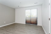 This single-floor unit offers a clean, contemporary design with a thoughtful layout and modern finishes throughout. The kitchen is a standout feature, equipped with stainless steel appliances including a refrigerator, electric range, and over-the-range microwave. Crisp white cabinetry with brushed nickel hardware extends to the ceiling, maximizing storage, while dark countertops provide a striking contrast. A kitchen island with an integrated sink and pendant lighting overhead adds both functionality and style. Light hardwood-style flooring runs through the main living and kitchen areas, contributing to a bright, cohesive aesthetic. The bedroom is finished with plush carpeting and features a generously sized window fitted with horizontal blinds, inviting natural light and offering views of surrounding greenery. The bathroom is clean and well-appointed, featuring light wood-style flooring, a full-size soaking tub with a shower and curved curtain rod, and a bright white finish throughout. The overall floor plan, as seen in the dollhouse view, reveals a compact yet well-organized layout with defined spaces for living, cooking, sleeping, and bathing — all presented in a neutral, move-in-ready palette.