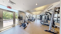 This fitness center offers a comprehensive workout environment housed within a single, open-plan floor. The space is thoughtfully designed with a neutral color palette of soft grays and whites, complemented by a durable speckled rubber flooring that provides both safety and comfort during exercise.

Large windows line the perimeter walls, flooding the room with abundant natural light and offering views of the surrounding outdoor greenery. The coffered ceiling features a combination of recessed lighting and suspended linear fixtures, ensuring even illumination throughout the space.

The cardio zone is well-appointed with multiple treadmills, elliptical trainers, stationary bikes — including a dedicated cycling bike — and a stair climber, several of which are equipped with personal monitors. The strength training area includes a multi-station cable and weight machine, a barbell rack with bench, and a dedicated dumbbell rack offering a range of free weights. Additional functional fitness accessories such as battle ropes and medicine balls are neatly organized along the walls.

A glass-paneled interior door connects the fitness center to an adjacent corridor, while a full-length glass exterior door provides direct outdoor access. Wall-mounted mirrors enhance the sense of space and allow users to monitor their form. The overall ambiance is clean, bright, and professional.
