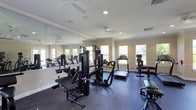 This single-level fitness room is designed to support a well-rounded workout routine, offering a thoughtfully arranged selection of exercise equipment within a bright, functional space. The room is outfitted with a variety of strength-training machines, including a multi-station cable system and a weight bench, alongside cardio equipment such as treadmills and a stationary bike with an integrated display screen.

The flooring is covered in dark rubber matting, providing durability and safety during exercise. A full-length mirror spans one wall, visually expanding the space and allowing users to monitor their form while training. Multiple windows along the adjacent wall invite generous natural light, contributing to an open and energizing atmosphere.

Crown molding along the upper perimeter adds a refined architectural detail, while the textured ceiling features recessed lighting and ceiling fans to ensure consistent illumination and air circulation throughout. The warm, neutral-toned walls complement the overall clean and functional aesthetic of the room, creating a comfortable environment suited for daily fitness use.