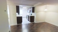 This single-floor space offers a clean, functional layout with a clear visual connection between the main living area and the kitchen. The living area is finished with rich, dark hardwood flooring that adds warmth and contrast to the neutral, light-toned walls. A subtle step-down or transition marks the boundary between the living space and the kitchen zone, where light-colored tile flooring takes over, creating a natural delineation between the two areas.

The kitchen is well-equipped and thoughtfully designed, featuring dark espresso-toned cabinetry both above and below the counters, providing ample storage. Light-colored countertops offer a crisp contrast to the cabinetry. Stainless steel appliances, including a range and microwave, are integrated seamlessly into the layout. A sink with a window above it allows for natural light to enter the kitchen. The ceiling in the kitchen features a drop-tile grid with recessed panel lighting, ensuring the space is well-lit. Baseboard heating is visible along the walls. Overall, the space presents a practical and tidy interior with a neutral palette and modern fixtures.