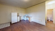 Located at 5324 E 1st St in Tucson, Arizona, this efficient single-floor studio-style unit offers a straightforward and functional layout. The main living area is an open, combined living and kitchen space finished with warm wood-style plank flooring throughout. The kitchen is equipped with a white refrigerator, an electric range, and a sink with adjacent counter space, all positioned along one wall to maximize the open feel of the room. A textured ceiling and light-colored concrete block walls give the space a utilitarian character. Natural light enters through a sliding window that frames a view of the surrounding neighborhood, including palm trees and adjacent structures, lending a touch of the Tucson landscape to the interior. A wall-mounted air conditioning unit provides climate control suited to the desert environment. The bathroom is a compact, fully tiled space featuring a vanity with under-sink storage, a toilet, and a combination tub and shower enclosed by a curtain. White subway-style tile lines the walls, maintaining a clean and simple aesthetic. A private entry door provides direct access to the unit. Overall, the space offers a no-frills, practical layout well-suited for single occupancy in a convenient Tucson location.