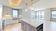 This well-appointed 2-bedroom, 1-bathroom residence offers a thoughtfully designed open-concept layout bathed in natural light. The living area is a standout, featuring dramatic floor-to-ceiling corner windows that frame sweeping panoramic city views, creating an airy, light-filled atmosphere throughout the day. Light hardwood flooring flows seamlessly through all main areas, lending warmth and continuity to the space.

The kitchen is a true centerpiece, showcasing a large island with striking blue cabinetry and white quartz countertops, complemented by soft gray upper cabinets with brass hardware. Globe pendant lights suspended from a coffered ceiling add an elegant finishing touch, while stainless steel appliances and a stainless farmhouse sink complete the functional design.

The generously sized primary bedroom offers ample space, a ceiling fan, and direct access to a beautifully appointed bathroom. The bathroom features floor-to-ceiling stone-look tile, a glass-enclosed soaking tub and shower combination, a white double vanity with brass fixtures, and sophisticated pendant lighting. The second bedroom is bright and comfortable, with a large window overlooking city rooftops. Both bedrooms are served by dedicated closets with adjustable shelving systems. A clean hallway with louvered closet doors connects the living spaces efficiently.