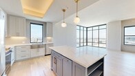 This well-appointed 2-bedroom, 1-bathroom residence offers a thoughtfully designed open-concept layout bathed in natural light. The living area is a standout, featuring dramatic floor-to-ceiling corner windows that frame sweeping panoramic city views, creating an airy, light-filled atmosphere throughout the day. Light hardwood flooring flows seamlessly through all main areas, lending warmth and continuity to the space.

The kitchen is a true centerpiece, showcasing a large island with striking blue cabinetry and white quartz countertops, complemented by soft gray upper cabinets with brass hardware. Globe pendant lights suspended from a coffered ceiling add an elegant finishing touch, while stainless steel appliances and a stainless farmhouse sink complete the functional design.

The generously sized primary bedroom offers ample space, a ceiling fan, and direct access to a beautifully appointed bathroom. The bathroom features floor-to-ceiling stone-look tile, a glass-enclosed soaking tub and shower combination, a white double vanity with brass fixtures, and sophisticated pendant lighting. The second bedroom is bright and comfortable, with a large window overlooking city rooftops. Both bedrooms are served by dedicated closets with adjustable shelving systems. A clean hallway with louvered closet doors connects the living spaces efficiently.