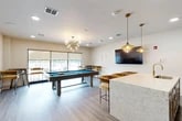 Located at 4781 SW 36 Rd in Gainesville, FL, this contemporary amenity lounge is a well-appointed recreational space designed for socializing and leisure. The open-plan layout is anchored by a full-size pool table with a light wood frame and vibrant blue felt surface, complemented by a wall-mounted cue rack and framed abstract artwork nearby. Overhead, a striking sputnik-style chandelier with globe bulbs serves as a visual centerpiece, while additional pendant lights in brushed gold finish illuminate a large quartz-topped bar counter with undermount sink and bar-height seating upholstered in warm textured fabric. A wall-mounted flat-screen TV provides an entertainment focal point, and the crisp white walls are further accented by a bold decorative wallpaper feature in the bar area. Light wood-look plank flooring runs throughout, lending warmth and continuity to the space. Expansive floor-to-ceiling windows with roller shades flood the lounge with natural light and offer direct views of an outdoor pool area complete with lounge seating. Recessed lighting complements the chandelier fixtures for a layered lighting design. The overall ambiance is modern, vibrant, and inviting — ideal for community gatherings and casual recreation.