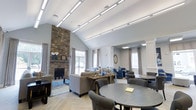 Located at 20 Hopmeadow in Aspen Glen, this single-story space offers a thoughtfully designed communal environment that blends rustic and contemporary elements. The most commanding architectural feature is the floor-to-ceiling stone fireplace, which anchors one end of the main room and adds a warm, grounding presence to the space.

The vaulted ceiling rises dramatically overhead, lined with sleek, modern linear lighting fixtures that complement the airy, open feel of the interior. Light-toned herringbone-patterned flooring runs throughout, contributing to the bright and cohesive aesthetic. Large windows dressed with elegant grey curtains allow generous amounts of natural light to filter in, enhancing the room's welcoming atmosphere.

The layout accommodates multiple seating arrangements, including plush sofas and armchairs grouped around a central coffee table, as well as round dining tables with upholstered chairs positioned closer to the foreground. A decorative arched passageway leads into an adjacent lounge area featuring additional seating and a framed mirror, creating a sense of depth and continuity throughout the space. Classic wainscoting panels add a refined architectural detail to the walls.