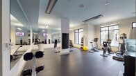 This fitness center is a thoughtfully designed, single-floor facility that offers a comprehensive workout environment for users of all fitness levels. The space features a clean, modern aesthetic with white walls, a light blue-toned ceiling, and durable rubber flooring throughout, providing both style and function.

Natural light pours in through multiple large windows along the perimeter walls, creating an open and energizing atmosphere. Full-length mirrors line one side of the facility, visually expanding the space and allowing users to monitor their form during exercise.

The cardio zone is well-appointed with stationary bikes, elliptical trainers, and treadmills positioned near the windows to take advantage of natural light and outside views. The strength training area includes a multi-station cable and weight machine, adjustable benches, and a dedicated free weights section featuring a rack of dumbbells and an assortment of medicine balls in varying weights.

Additional amenities include wall-mounted flat-screen televisions for entertainment during workouts and a water cooler station for hydration convenience. Structural columns are integrated throughout the layout, lending an architectural character to the space without impeding workflow. The facility is accessed through a set of glass-paneled doors, contributing to the overall transparency and modern design of this versatile fitness space.