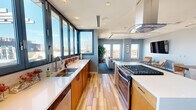 Located at 100 Steele St in Denver's Cherry Creek neighborhood, this contemporary single-floor space offers a thoughtfully designed open-plan layout that seamlessly connects the kitchen, dining, and lounge areas. The fully equipped kitchen is a standout feature, boasting warm wood cabinetry, white quartz countertops, a large center island with a gas cooktop and stainless steel hood, a full-size refrigerator, and a wall of floor-to-ceiling windows that flood the space with natural light and frame sweeping outdoor views. The dining area accommodates generous seating around a rustic solid-wood table paired with modern chrome-and-leather chairs, with blue abstract artwork adorning the soft gray walls. The lounge area features plush seating, a large wall-mounted display, and patterned carpet underfoot, creating a relaxed yet refined atmosphere. Double glass doors open directly to an exterior terrace, further enhancing the connection to the outdoors and offering elevated views of the Denver skyline. Throughout the space, wood-look flooring, recessed lighting, and a cohesive cool-gray palette contribute to a modern, professional, and welcoming ambiance.