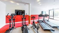 Located at 8740 S La Tijera Blvd in Los Angeles, this single-floor fitness center is designed to support a comprehensive workout experience. The space is defined by a clean, energetic aesthetic, with white upper walls accented by bold red and orange lower panels that create a motivating atmosphere throughout.

The cardio zone is well-stocked with multiple treadmills, elliptical trainers, and stationary cycling bikes, all positioned to face wall-mounted flat-screen televisions for an engaging workout experience. Strength training is supported by a multi-functional cable machine and a dedicated resistance equipment station, offering a variety of exercise options for users of all fitness levels.

A large wall mirror expands the perceived depth of the room while allowing users to monitor their form during exercise. Recessed ceiling lighting provides consistent, even illumination across the space. A glass door along one wall introduces natural light and opens to an outdoor area, adding a sense of openness to the interior. The flooring transitions between cushioned neutral carpet and durable red rubber matting, delineating activity zones effectively. Double entry doors with a branded entry mat complete the functional, polished design of the facility.