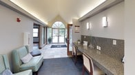 This single-floor community amenity building offers a thoughtfully designed space that balances functionality with an inviting atmosphere. The entrance lobby immediately impresses with its soaring vaulted ceiling and a striking arched transom window above the double-door entry, flooding the space with natural light and providing views of the surrounding exterior grounds. Stone-tiled flooring transitions seamlessly into plush blue carpeting, grounding the space with a clean, polished aesthetic.

The reception area features a generously sized granite-topped counter with built-in seating, accented by elegant wall-mounted sconce lighting in a chrome finish. Informational displays and decorative touches, including a glass vase with greenery and small potted plants, lend a welcoming, professional character to the check-in station.

Adjacent to the entry, a comfortable lounge area offers soft seating with light sage-green upholstered chairs and decorative pillows, perfect for relaxed conversation or waiting. Cove lighting along the upper walls adds a warm, ambient glow throughout.

The fitness center is accessed through full-height wood-framed glass doors, offering excellent visibility into the well-equipped space. Inside, a variety of cardio machines and exercise equipment are arranged across a rubberized floor, with mirrored walls and windows providing natural light. A beverage vending machine is conveniently positioned just outside the fitness room entrance, completing this practical and well-appointed facility.