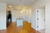 This single-floor residence showcases a thoughtfully designed open-concept layout where the kitchen flows seamlessly into the living area. The kitchen serves as the centerpiece of the space, featuring a large central island topped with dark granite countertops and equipped with a gold-tone faucet and undermount sink. Pendant lights with glass shades hang above the island, adding a contemporary touch. Slate-blue shaker-style cabinetry lines the kitchen walls, complemented by a herringbone tile backsplash and a full suite of stainless steel appliances, including a range, microwave, and refrigerator.

Hardwood-style flooring extends throughout the main living areas, offering a warm contrast to the crisp white walls. Natural light enters through several windows, brightening the open living space, which is finished with a ceiling fan overhead. The bedroom is carpeted and similarly well-lit, with a ceiling fan and a window overlooking the outdoors.

The bathroom features a dual-sink vanity with matching slate-blue cabinetry, granite countertops, gold hardware and fixtures, a large framed mirror, and a Hollywood-style light bar, creating a polished and cohesive aesthetic throughout the unit.