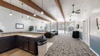 This single-floor community space offers an open and airy layout that seamlessly integrates a kitchen, dining area, and living room into one cohesive environment. The vaulted ceiling is a defining architectural feature, adorned with bold exposed wooden beams that run the length of the room, adding warmth and a rustic character to the otherwise contemporary interior. Multiple ceiling fans are suspended throughout, ensuring comfortable airflow across the generously sized floor plan.

The kitchen area is anchored by a striking blue-accented breakfast bar with a granite-style countertop and bar stools, complemented by dark espresso cabinetry, a sink, and built-in appliances including a microwave and coffee station. A standout adobe-style kiva fireplace with a brick interior adds a distinctive southwestern architectural touch adjacent to the kitchen.

The living area is carpeted in a patterned, neutral-toned tile-effect carpet and is furnished with comfortable seating. A wall-mounted television provides entertainment, while a reception-style dark cabinet unit serves as a media console. The dining area features a bistro-style glass table positioned near floor-to-ceiling sliding glass doors and windows that flood the space with natural light and offer direct views of a lush outdoor pool courtyard. French doors on either side provide additional outdoor access, enhancing the indoor-outdoor connection throughout this welcoming communal space.