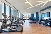 Located at 201 SE Sixth Ave in Fort Lauderdale, this premium fitness center offers a generously proportioned, single-floor facility designed to support a comprehensive range of workout disciplines. The main gym floor is defined by warm hardwood-style flooring, textured blue-gray accent walls, and striking geometric linear lighting fixtures overhead that lend the space a modern, upscale aesthetic. Floor-to-ceiling windows wrap around the perimeter, flooding the interior with abundant natural light and framing elevated views of the Fort Lauderdale skyline.

The main workout area is comprehensively equipped with cardio machines including treadmills, ellipticals, and stair climbers, alongside an extensive free weight section featuring a full dumbbell rack, adjustable benches, cable machines, and functional training rigs with medicine balls and kettlebells. Mirrored wall panels are strategically placed throughout to enhance the sense of space and support form monitoring.

A dedicated Pilates and group fitness studio features reformer machines, stability balls, resistance accessories, and a vibrant tropical-themed accent wall, enclosed by sleek black-framed glass partitions. Adjacent to it, a separate spin studio houses multiple cycling bikes with integrated screens and benefits from its own window views. The overall environment is polished, well-lit, and purposefully organized for peak performance.