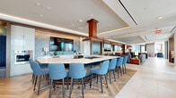 Located at 300 K St in Washington, DC, this generously sized amenity lounge spans a single floor and offers a sophisticated blend of social, dining, and collaborative spaces. The open-plan interior is anchored by a large kitchen island with bar seating, a statement copper range hood overhead, and a full suite of appliances — ideal for gatherings and entertaining. Adjacent lounge areas feature carefully curated furniture arrangements, including plush sofas, accent chairs, and a large-screen media wall framed by warm wood paneling and bold geometric detailing, all set on a combination of herringbone hardwood and large-format tile flooring. Cove lighting and recessed fixtures throughout create a warm, inviting ambiance. A dedicated conference room with floor-to-ceiling windows, an oval wood table, and dramatic pendant lighting offers a more focused setting. The space connects seamlessly to multiple outdoor terraces, where wood-deck flooring, teal outdoor seating, hanging egg chairs, and ceiling fans make the most of open-air Washington views. Natural light pours in through expansive glass doors and windows, blurring the line between interior comfort and outdoor living. The overall design is modern, polished, and thoughtfully appointed for both relaxation and productivity.