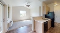 This single-floor residence offers a thoughtfully designed open layout with a seamless flow between living and kitchen spaces. The living area is a standout, boasting soaring vaulted ceilings fitted with a ceiling fan, neutral carpet flooring, and a large grid-pane window that floods the room with natural light and frames views of surrounding trees. Hardwood-style flooring transitions the space into the kitchen, which features light wood cabinetry, white countertops, and a full suite of black appliances including a range with microwave, refrigerator, and dishwasher. A peninsula with a built-in sink separates the kitchen from the living area, creating a functional and social cooking space. The galley-style kitchen corridor leads conveniently to the entry door. The bedroom is generously proportioned with plush carpeting and a large window overlooking lush greenery, providing a calm, private retreat. The bathroom is clean and modern, outfitted with light wood cabinetry, white countertops, brushed nickel hardware, and a separate area with a towel bar and natural light from an upper window. Throughout, the unit maintains a consistent palette of soft whites and warm wood tones, creating a welcoming and contemporary ambiance.