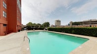 This outdoor pool area offers a refreshing retreat set against a backdrop of an urban skyline. The generously sized swimming pool features clear turquoise water, with entry points provided by metal handrails on multiple sides, making it accessible and convenient for swimmers. The surrounding concrete deck provides ample space for lounging and movement, and is furnished with reclining lounge chairs positioned to take advantage of the open-air environment.

The perimeter of the pool area is lined with well-maintained hedging and wooden fencing, offering a degree of privacy while still allowing views of the surrounding cityscape, including notable mid-rise and high-rise buildings visible on the horizon. A small grassy area with mature trees adds a natural element to the otherwise paved setting, along with potted planters that contribute greenery to the space.

Practical amenities include waste receptacles placed throughout the deck area. Access to the adjacent building is provided via a secured glass door entrance, posted with a no-smoking policy. The area is open to the sky, allowing abundant natural light throughout the day and creating a bright, inviting atmosphere suitable for outdoor recreation.