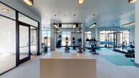 This single-floor fitness center is thoughtfully designed to accommodate a wide range of workout needs within a spacious, open layout. The facility is divided into distinct zones, each serving a specific training purpose while maintaining a cohesive, contemporary aesthetic throughout.

The main cardio and strength training area is the largest section of the facility, featuring an extensive collection of equipment including treadmills, ellipticals, stair climbers, rowing machines, and stationary bikes. Exposed concrete columns punctuate the space, while a combination of pendant lighting and decorative ceiling-mounted fixtures provide ample illumination. Large, floor-to-ceiling windows line the perimeter, flooding the area with natural light and offering open views of the surrounding exterior. Ceiling fans contribute to comfortable airflow throughout.

A dedicated strength training zone houses a power rack with a wooden platform, weight benches, cable machines, and Precor selectorized equipment, all positioned on teal-colored rubber flooring that defines the area.

A separate studio room features warm wood flooring, a dark-painted ceiling, full-wall mirrors, built-in open shelving, and open floor space suited for yoga, cycling, or group fitness. The reception area near the entrance includes a central storage island and decorative pendant lighting, providing a welcoming transition into the facility.