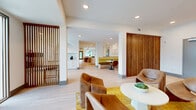 This expansive single-floor amenity space presents a thoughtfully designed open layout that seamlessly blends mid-century modern aesthetics with contemporary finishes. Light oak hardwood flooring runs throughout, complementing crisp white walls and recessed lighting that keep the environment bright and welcoming.

The space is organized into several functional zones. A welcoming entry lounge near the main glass doors features a cluster of cognac leather lounge chairs arranged around a round white pedestal table, anchored by a bold yellow patterned area rug. Vertical wood slat panel dividers add warmth and visual rhythm, subtly defining zones without sacrificing openness.

Moving deeper into the space, a central gallery corridor is lined with striking large-format artwork, including a vibrant tropical leaf painting and textured wood sculptural wall pieces. A kitchenette area with white cabinetry and counter seating offers utility, while a built-in booth with an olive-green banquette provides an intimate dining nook. A live-edge communal table with bench seating anchors the main gathering area.

Toward the far end, a quieter lounge alcove features sage green accent chairs, a natural wood round coffee table, and a large picture window framing lush palm tree views, filling the corner with natural light and a serene outdoor connection.