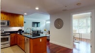 This single-floor residence offers a thoughtfully designed layout that balances open communal spaces with private retreats. Rich cherry hardwood flooring flows throughout the main living areas, creating a warm and cohesive aesthetic. The open-concept kitchen is a standout feature, equipped with stainless steel appliances including a gas range, built-in microwave, and dishwasher, all complemented by warm wood cabinetry and dark granite countertops. A central island with a sink adds both functionality and a natural gathering point. The kitchen opens seamlessly into the living area, where a wall-mounted flat-screen TV and comfortable sectional seating create an inviting space for relaxation. A dedicated dining area accommodates a glass-top table with modern white chairs, framed by abstract artwork and a large indoor plant. The bedroom is bright and serene, benefiting from multiple windows that invite natural light and views of surrounding greenery, with ample space for a full furniture arrangement. The bathroom features marble-tiled walls and flooring, a combination tub and shower, a white vanity with stone countertop, and a large mirror. The entry hallway provides practical storage and connects all areas of the residence with a clean, neutral palette throughout.
