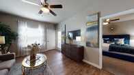 This single-floor residence offers a cohesive and thoughtfully designed living space with a warm, contemporary aesthetic throughout. Rich hardwood-style flooring flows through the main living areas, complemented by neutral gray and taupe wall tones that create a calm, inviting atmosphere.

The living room is a comfortable and well-lit space, featuring a ceiling fan with integrated lighting and large windows dressed with sheer, patterned curtains that allow natural light to filter in. A cozy seating arrangement anchors the room, and a compact dining nook with a bistro-style table and acrylic chairs sits conveniently near the entry door.

The kitchen is well-equipped with dark espresso cabinetry, light granite-style countertops, a tiled backsplash, and a breakfast bar peninsula that provides additional prep and casual dining space. Hardwood-style flooring continues here, tying the space together seamlessly.

The bedroom is carpeted for comfort and features a ceiling fan, a closet with rod storage, and a window with patterned drapes. It connects openly to the living area, enhancing the sense of flow throughout the residence.

The bathroom includes dark cabinetry with ample under-sink storage, a granite-style vanity countertop, a large wall mirror, and additional upper cabinetry for storage. Overall, the space presents a balanced combination of function and modern style.