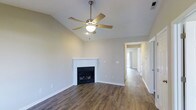 This single-story residence offers a thoughtfully designed layout with a warm, inviting ambiance. The main living area immediately draws attention with its vaulted ceilings that lend a sense of spaciousness and openness to the room. A ceiling fan with a built-in light fixture is centrally mounted, providing both comfort and illumination. A classic white-mantled fireplace serves as the focal point of the living space, adding charm and a cozy atmosphere. Rich, dark hardwood-style flooring runs throughout, providing a stylish and durable foundation that complements the neutral wall tones. The soft, light gray walls create a clean and versatile backdrop suitable for a variety of décor styles. A hallway extends from the living area, connecting to additional rooms and hinting at a well-organized, flowing floor plan. Natural light filters through windows visible further down the corridor, brightening the interior. The overall design balances practicality with comfort, making it a functional and welcoming single-floor living space.