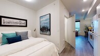 This thoughtfully designed single-floor residence offers a cohesive and contemporary layout with clean lines and a neutral color palette throughout. The living area serves as the heart of the space, featuring warm wood-look flooring, a ceiling fan, and a striking large-format artwork above the sofa. A floor-length mirror and area rug add texture and depth to the room. The standout feature of the living area is the expansive floor-to-ceiling corner window that floods the space with natural light and offers sweeping views of the surrounding neighborhood and treeline, creating an airy, open ambiance. A dedicated workspace with a desk and task lighting is neatly integrated into this area. The bedroom is calm and well-proportioned, with crisp white walls, wood-look flooring, and curated artwork including a bold cityscape painting. A brass bedside lamp adds a warm accent. The bathroom is finished in a clean, minimalist style with white cabinetry, an under-mount sink, white countertops, a large mirror, and wood-look flooring that ties back to the rest of the unit. The overall design is modern and cohesive, balancing comfort and style with practical, functional layouts throughout.