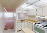 Located at 1400 NW Tenth Ave in Miami, FL, this single-floor unit offers a straightforward layout designed for practical living. The kitchen is equipped with white cabinetry providing ample storage, light-toned laminate countertops arranged in an L-shaped configuration, a double basin sink, an electric range with overhead exhaust, and a full-size refrigerator. A drop-panel ceiling with built-in fluorescent lighting illuminates the kitchen workspace, while tile flooring adds easy-clean durability underfoot. A pass-through opening connects the kitchen to the adjacent hallway, which transitions into the carpeted living area.

The living space features warm-toned carpet flooring, a ceiling fan, and a textured ceiling throughout. The bedroom area is generously proportioned, bathed in natural light from a large window fitted with vertical blinds that frame views of the surrounding Miami skyline and greenery. The neutral white walls and soft carpet create a calm, functional environment.

The bathroom includes a vanity with a white basin, under-sink cabinetry, framed mirrors, a Hollywood-style light bar, and mosaic tile wainscoting along the walls, lending a clean and classic finish. Overall, the unit presents a clean, functional aesthetic suited to urban Miami living.