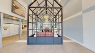 Located at 1230 Vintage Ln in Morgan Hill, California, this single-story community clubhouse is a striking example of contemporary design. The centerpiece of the space is a bold glass-and-steel greenhouse-style atrium structure that rises to soaring vaulted ceilings, anchoring the main lounge area with architectural drama. Exposed dark steel truss framing contrasts beautifully against bright white walls and an abundance of natural light that pours in through expansive windows and the glass ceiling panels above.

The lounge area is elegantly appointed with a vibrant plaid sofa, sculptural accent stools, layered area rugs, and carefully curated artwork, creating a warm yet sophisticated atmosphere. Large-format tile flooring in a soft stone tone flows throughout the main entry and common areas, while wood-look flooring adds warmth in adjoining spaces.

The entry foyer makes a memorable first impression with a textured tile accent wall, wall-mounted artwork, bench seating, and floor-to-ceiling windows that invite natural light. Adjacent to the main lounge, a dedicated package room with secure lockers provides added convenience. Additional rooms visible from the central space include a stylishly finished kitchenette or service area with light wood cabinetry and modern fixtures. Glass partitions and doors connect the various zones while maintaining an open, airy feel throughout.