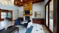 Located at 6310 Ansley Falls Dr in Charlotte, NC, this sophisticated single-floor community space showcases a refined blend of natural materials and thoughtful design. The main lounge area is anchored by a striking floor-to-ceiling stone fireplace with a wood mantel, above which a mounted flat-screen television is seamlessly integrated. Warm, coffered wood ceilings with recessed lighting lend a sense of height and architectural distinction to the interior.

The flooring throughout is composed of elegant stone tile laid in a classic pattern, accented with dark granite inlays that add visual interest and depth. Generous windows with rich wood trim frame views of lush greenery, filling the space with abundant natural light. French doors open directly to an outdoor covered patio area, creating a seamless indoor-outdoor connection.

The lounge is furnished with comfortable navy blue seating, including a curved sofa and accent chairs, arranged around a dark wood coffee table atop a decorative area rug. Dark cherry wood cabinetry with granite countertops provides additional storage and display surfaces along the perimeter walls. A contemporary bubble chandelier adds a modern contrast to the traditional wood tones. The overall ambiance is warm, inviting, and well-appointed, ideal for gathering and relaxation.