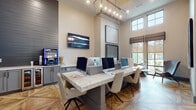 Located at 8050 Lapis Ln NW in Concord, NC, this stylish single-floor community clubhouse blends contemporary design with functional amenities throughout its thoughtfully curated spaces. The interior is unified by striking geometric-patterned hardwood flooring that adds warmth and visual interest underfoot, while soaring ceilings and tall, multi-pane windows flood the space with natural light and outdoor views.

The business center serves as a productive hub, featuring a generous marble-topped communal work surface lined with Apple iMac workstations, complemented by a modern linear chandelier overhead and wall-mounted display screens. Curated abstract artwork adorns the walls, contributing to the refined, professional atmosphere.

A well-appointed kitchenette offers grey shaker-style cabinetry, marble-look countertops, a sink, open rustic wood shelving against a teal shiplap accent wall, and a beverage cooler — ideal for hosting and everyday convenience.

Dramatic floor-to-ceiling steel-framed glass partition walls define zones while preserving an open, connected feel throughout. A comfortable lounge area with mid-century modern accent chairs and a marble side table provides a welcoming spot for relaxation beside floor-to-ceiling windows. Stone accent walls and arched architectural details add character, making this clubhouse an inviting and functional gathering destination.