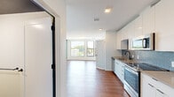 Located at 221 S Huntington Ave in Boston's Jamaica Plain neighborhood, this efficiently designed unit offers a thoughtful open-concept layout that combines the kitchen and living area into one cohesive space. Rich hardwood flooring runs throughout, anchoring the bright, contemporary aesthetic defined by crisp white walls and recessed lighting.

The kitchen is a standout feature, outfitted with sleek white flat-front cabinetry extending to the ceiling for ample storage, a striking blue geometric tile backsplash, quartz countertops, and a full suite of stainless steel appliances including a range, microwave, dishwasher, and refrigerator. A matte black faucet adds a refined contrast to the overall design.

The open living area benefits from a wall of windows that floods the space with natural light and provides treetop views of the surrounding neighborhood. A teal accent wall adds a bold, modern touch to the otherwise neutral palette.

The bathroom is well-appointed with hexagonal floor tile in a warm taupe tone, white subway-tiled shower walls surrounding a full soaking tub, matte black fixtures, and a warm wood vanity. Clean lines and quality finishes are consistent throughout, making this a polished and functional urban residence.