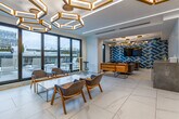 Located at 50 Portland Street in Toronto's vibrant King West neighbourhood, this thoughtfully designed communal space combines a fully equipped kitchen with an expansive open-plan living area. The interior immediately impresses with its signature ceiling installation — a series of large, gold-framed hexagonal light fixtures that cast a warm, ambient glow across the entire floor. Large-format white marble-effect porcelain tiles stretch across the floor, lending a clean and polished foundation to the space. Floor-to-ceiling windows and glass sliding doors line the perimeter, flooding the interior with natural light and offering views of the surrounding cityscape and an adjacent outdoor terrace. The kitchen is a standout feature, showcasing warm walnut cabinetry, a concrete-finish island with bar-height seating, stainless steel appliances, and a dramatic geometric-patterned backsplash in gold, black, and white tones. The living area is divided into multiple distinct seating zones, each furnished with contemporary sofas and lounge chairs in complementary muted tones. A bold blue geometric accent wall adds a striking focal point to one corner of the room. The overall ambiance is modern, refined, and inviting — a space designed for both productivity and relaxation.