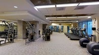 This fitness center is designed to accommodate a comprehensive workout experience within a single, open-plan floor. The space is generously sized, offering ample room to move freely between equipment stations without feeling crowded. A row of treadmills lines one side of the facility, providing dedicated space for cardiovascular training, while a variety of cable and weight machines are arranged throughout the strength training area. Concrete columns punctuate the space, adding an industrial character that contrasts with the neutral, light-toned carpet flooring underfoot. The dropped ceiling features a grid of bright fluorescent panel lighting, ensuring the entire area is evenly and thoroughly illuminated. Large mirrors are mounted along the walls, visually expanding the space and allowing users to monitor their form during exercise. The overall ambiance is clean, functional, and professional, reflecting the design priorities of a well-maintained commercial fitness facility. The layout encourages an efficient flow from one equipment zone to another, making the space practical for users with varied training goals.