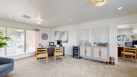 This single-floor office space combines functionality with a warm, inviting aesthetic throughout its thoughtfully designed layout. The main workspace features a substantial dark wood executive desk accompanied by guest seating, a computer workstation, and framed abstract artwork displayed on crisp white walls. Recessed lighting and elegant woven pendant light fixtures illuminate the space, while white plantation-shuttered windows allow ample natural light to filter in. A beverage refrigerator stocked with bottled water sits beside a stylish gray cane-paneled credenza, which serves as a refreshment station complete with coffee amenities and neatly arranged baskets. An adjacent workspace area includes a dark wood lateral filing cabinet and a multifunction printer, providing a practical support zone for daily operations. The reception lounge area offers a relaxed seating arrangement with plush blue accent chairs, a wooden open-shelf display unit adorned with woven wall décor, and lush indoor plants that add a natural, welcoming touch. Soft peach curtains frame the glass-paned exterior door and surrounding windows, offering views of the outdoor greenery beyond. Neutral patterned carpeting flows throughout the space, tying together the cohesive, contemporary design that balances professionalism with comfort.