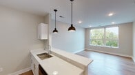 This single-floor unit offers a clean, contemporary design characterized by warm wood-look flooring, neutral wall tones, and modern finishes throughout. The open-concept layout connects the kitchen to the living area, creating a cohesive and functional flow. The kitchen is well-appointed with white shaker-style cabinetry, stainless steel appliances including a range, microwave, and refrigerator, a subway tile backsplash, and light quartz countertops that extend to a peninsula with an undermount sink. Two bronze dome pendant lights hang above the peninsula, adding a stylish accent. The living area benefits from natural light through a large picture window with views of surrounding greenery and neighboring architecture, complemented by recessed ceiling lighting. The bedroom is a comfortable, well-proportioned space featuring a ceiling fan with integrated lighting, a window allowing natural light, and the same warm flooring found throughout. The bathroom includes a white vanity with a vessel-style sink, light quartz countertop, a large framed mirror, and a shower with a curtain rod. Louvered closet doors provide additional storage. Throughout the unit, clean white baseboards and neutral finishes contribute to a bright, move-in-ready atmosphere.