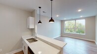This single-floor unit offers a clean, contemporary design characterized by warm wood-look flooring, neutral wall tones, and modern finishes throughout. The open-concept layout connects the kitchen to the living area, creating a cohesive and functional flow. The kitchen is well-appointed with white shaker-style cabinetry, stainless steel appliances including a range, microwave, and refrigerator, a subway tile backsplash, and light quartz countertops that extend to a peninsula with an undermount sink. Two bronze dome pendant lights hang above the peninsula, adding a stylish accent. The living area benefits from natural light through a large picture window with views of surrounding greenery and neighboring architecture, complemented by recessed ceiling lighting. The bedroom is a comfortable, well-proportioned space featuring a ceiling fan with integrated lighting, a window allowing natural light, and the same warm flooring found throughout. The bathroom includes a white vanity with a vessel-style sink, light quartz countertop, a large framed mirror, and a shower with a curtain rod. Louvered closet doors provide additional storage. Throughout the unit, clean white baseboards and neutral finishes contribute to a bright, move-in-ready atmosphere.