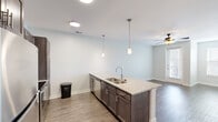 This single-floor unit offers a clean, modern aesthetic defined by a neutral color palette, wood-look plank flooring, and crisp white walls throughout. The open-concept layout connects the kitchen and living area seamlessly, creating a spacious and functional flow. The kitchen features a peninsula island with granite countertops, a stainless steel undermount sink, and dark espresso cabinetry with brushed nickel hardware. Pendant lights hang above the island, complementing the recessed lighting overhead. Ample upper and lower cabinetry, including a tall pantry-style unit, provides generous storage. The living area benefits from natural light through a window and is equipped with a ceiling fan. A glass-panel exterior door in one area allows additional natural light to filter in while providing outdoor access. The bathroom is finished consistently with the rest of the unit, featuring granite-topped dark cabinetry, a tub-shower combination, and wood-look flooring. Additional rooms are bright and open, with panel-style interior doors and well-proportioned layouts suited for flexible use. Overall, the unit presents a cohesive, contemporary finish throughout.