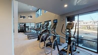 This fitness center is designed to provide a comprehensive workout experience within a spacious, modern environment. The facility features dramatically high ceilings that amplify the sense of openness throughout the space. Large floor-to-ceiling windows line the walls, flooding the interior with natural light and offering views of the surrounding landscaped outdoor areas, creating an energizing atmosphere for exercise.

The cardio zone is prominently equipped with multiple Matrix-brand treadmills, elliptical trainers, and stair climbers, each outfitted with individual display screens for entertainment during workouts. The equipment is arranged in organized rows along the windowed walls, maximizing both space and views.

A dedicated strength-training area features a cable machine, multi-function weight apparatus, and a dumbbell rack, providing a full range of resistance training options. The open floor area between zones offers ample room for stretching or functional training.

Flooring transitions between neutral-toned carpet and darker patterned sections help define workout zones throughout the space. Steel-framed glass partitions add an industrial-modern aesthetic detail. Recessed lighting and decorative artwork on the walls contribute to the polished, contemporary ambiance of the overall facility.