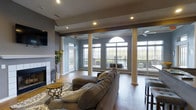 This single-floor space is a well-appointed communal lounge area designed to offer both comfort and sophistication. The open-plan layout is anchored by a row of tall, decorative columns that lend a sense of architectural elegance while defining the space without enclosing it. Warm hardwood flooring runs throughout, complementing the cool, neutral blue-gray tones of the walls and creating a balanced, inviting ambiance.

A classic white fireplace with a tiled surround serves as a focal point on one end of the room, adding a cozy element to the otherwise modern setting. Plush, sectional seating is arranged around a patterned area rug, providing ample room to relax and socialize. Ceiling fans are installed throughout to ensure comfortable air circulation.

Natural light pours in abundantly through an impressive array of large windows and a distinctive arched transom window, offering panoramic views of an outdoor pool and patio area. Glass-panel doors provide direct access to the exterior, further blurring the line between indoors and outdoors. Recessed lighting supplements the natural light, ensuring the space remains bright and welcoming at all hours. A wall-mounted television adds a practical amenity to this thoughtfully designed communal gathering area.