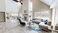 This single-floor space makes a striking first impression with its dramatic vaulted ceilings supported by exposed dark wood beams, creating an airy, open atmosphere throughout. The expansive living room serves as the centerpiece, anchored by a floor-to-ceiling shiplap fireplace that draws the eye upward and adds a refined, transitional architectural detail. Light wood-tone tile flooring flows seamlessly across the entire space, lending a clean, cohesive aesthetic.

An abundance of oversized windows on multiple walls frames picturesque wooded winter views, flooding the interior with natural light and creating a strong connection to the outdoors. The living area comfortably accommodates a large sectional seating arrangement, complemented by a statement area rug in blue and grey tones that adds warmth and texture.

An adjacent dining area with bar-height seating sits beneath the cathedral ceiling, seamlessly integrated into the open floor plan. A coffee bar area with dark cabinetry, a beverage cooler, and counter seating provides a functional and welcoming hospitality element. A glass-paneled entry with a full-view door connects the space to the exterior. A wall-mounted display screen and thoughtfully placed accent lighting round out this well-appointed, versatile space ideal for gathering and entertaining.