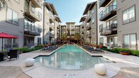 Located at 5940 Forest Park Rd in Dallas, Texas, this outdoor amenity space serves as the centerpiece of a modern multi-story residential community. The courtyard is anchored by a generously proportioned resort-style swimming pool with crystal-clear blue water, flanked on both sides by rows of sleek black chaise lounges accented with bold red cushions, providing ample space for relaxation and recreation. A shallow wading area and a separate spa are integrated into the pool design, offering varied aquatic experiences. The surrounding deck features a polished concrete surface with sculptural white accent pieces that add a contemporary touch. Tall palm trees and carefully maintained greenery lend a tropical ambiance to the outdoor space. The courtyard is enclosed by the multi-story residential building, whose exterior combines neutral gray siding with large windows and private balconies, creating an intimate yet open atmosphere. A covered pavilion area is visible at the far end of the courtyard, providing a shaded gathering spot. Wrought iron fencing with gated access defines the perimeter, ensuring security and privacy. Open to the sky, the space benefits from abundant natural light throughout the day, making it a vibrant outdoor retreat within the heart of Dallas.