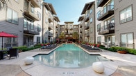 Located at 5940 Forest Park Rd in Dallas, Texas, this outdoor amenity space serves as the centerpiece of a modern multi-story residential community. The courtyard is anchored by a generously proportioned resort-style swimming pool with crystal-clear blue water, flanked on both sides by rows of sleek black chaise lounges accented with bold red cushions, providing ample space for relaxation and recreation. A shallow wading area and a separate spa are integrated into the pool design, offering varied aquatic experiences. The surrounding deck features a polished concrete surface with sculptural white accent pieces that add a contemporary touch. Tall palm trees and carefully maintained greenery lend a tropical ambiance to the outdoor space. The courtyard is enclosed by the multi-story residential building, whose exterior combines neutral gray siding with large windows and private balconies, creating an intimate yet open atmosphere. A covered pavilion area is visible at the far end of the courtyard, providing a shaded gathering spot. Wrought iron fencing with gated access defines the perimeter, ensuring security and privacy. Open to the sky, the space benefits from abundant natural light throughout the day, making it a vibrant outdoor retreat within the heart of Dallas.