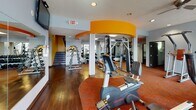 Located at 1344 Emajean St in Kalamazoo, Michigan, this single-level fitness center offers a comprehensive workout environment designed to accommodate a variety of exercise needs. The space is visually defined by bold orange architectural accents—including curved ceiling fascias and accent walls—set against neutral gray walls, creating an energetic and motivating atmosphere throughout.

The cardio zone is well-stocked with multiple treadmills, elliptical trainers, stair climbers, and stationary bikes, all arranged on richly patterned red-orange carpeting. Ceiling fans and recessed lighting provide comfort and visibility, while wall-mounted televisions offer entertainment during workouts. Large windows along one wall allow generous natural light to fill the space and provide views of the surrounding area.

The strength training section features a multi-station cable machine, free weights, weight plates, and benches positioned on hardwood flooring, providing a sturdy surface for resistance exercises. Full-length mirrors are strategically placed throughout to assist with form and to visually expand the space.

A dedicated functional training and stretching area includes exercise balls mounted on a vibrant orange wall, resistance bands, towel bars, stacked floor mats, and rubber flooring. A Western Michigan University Broncos mural adds local character and school spirit to the overall design.
