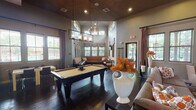 Located at 8601 Ice House Dr in North Richland Hills, TX, this single-floor community clubhouse is designed for socializing and relaxation. The space showcases soaring ceilings with sophisticated recessed lighting and elegant pendant chandeliers that illuminate rich, dark hardwood flooring throughout. The central game room is anchored by a full-size pool table beneath a striking multi-tier chandelier, flanked by plush leather seating and decorative accents that add warmth and character. Adjacent to the game area, a cozy lounge features a large flat-screen TV, a stylish credenza, and an eclectic mix of upholstered seating arranged over a bold patterned area rug. A tucked-away banquette nook, lined with built-in leather bench seating and wrap-around windows, offers views of the outdoor pool area and serves as a quiet retreat. The kitchen and dining zone is outfitted with a granite-topped bar counter, modern white bar stools, industrial-style pendant lighting with a vibrant orange ceiling accent, a full-size refrigerator, and nearby dining tables. Multiple windows throughout the clubhouse invite abundant natural light and outdoor views, while the cohesive warm-toned color palette, rich textiles, and curated décor create an inviting, upscale atmosphere ideal for community gatherings.
