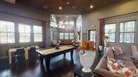Located at 8601 Ice House Dr in North Richland Hills, TX, this single-floor community clubhouse is designed for socializing and relaxation. The space showcases soaring ceilings with sophisticated recessed lighting and elegant pendant chandeliers that illuminate rich, dark hardwood flooring throughout. The central game room is anchored by a full-size pool table beneath a striking multi-tier chandelier, flanked by plush leather seating and decorative accents that add warmth and character. Adjacent to the game area, a cozy lounge features a large flat-screen TV, a stylish credenza, and an eclectic mix of upholstered seating arranged over a bold patterned area rug. A tucked-away banquette nook, lined with built-in leather bench seating and wrap-around windows, offers views of the outdoor pool area and serves as a quiet retreat. The kitchen and dining zone is outfitted with a granite-topped bar counter, modern white bar stools, industrial-style pendant lighting with a vibrant orange ceiling accent, a full-size refrigerator, and nearby dining tables. Multiple windows throughout the clubhouse invite abundant natural light and outdoor views, while the cohesive warm-toned color palette, rich textiles, and curated décor create an inviting, upscale atmosphere ideal for community gatherings.