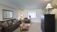 This single-floor residential space offers a clean, comfortable living environment with a thoughtfully arranged layout. The living room serves as the heart of the space, featuring wall-to-wall carpeting in a warm neutral tone that adds softness underfoot. Smooth, light-colored walls provide a bright and airy backdrop throughout, complemented by a ceiling with gentle architectural variation that adds subtle visual interest. A centrally positioned window allows natural light to fill the room, creating a welcoming atmosphere. The living area comfortably accommodates a full seating arrangement, including a plush sofa and an accent armchair, centered around a round wooden coffee table — ideal for relaxed everyday living or casual entertaining. A tall, dark-finished armoire anchors one side of the room, offering practical storage while contributing a grounded, classic aesthetic. Warm ambient lighting from a table lamp adds a cozy feel during evening hours. The open flow of the space allows for flexible furniture placement and makes the overall area feel generous and uncluttered. The neutral color palette throughout makes the space highly adaptable to a variety of décor styles, offering a versatile and inviting residential environment.