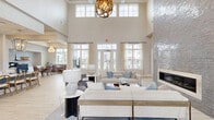 Located at 420 Angler Dr in Wilmington, NC, this generously sized single-floor community space offers a sophisticated coastal aesthetic with thoughtfully designed zones for gathering, lounging, and dining. The centerpiece of the main living area is a floor-to-ceiling tiled fireplace surround with a sleek linear firebox, flanked by soaring double-height ceilings adorned with elegant chandelier lighting and rows of transom windows that flood the space with natural light. French doors open to exterior views, enhancing the bright and airy ambiance. Light wood-look flooring runs throughout, complementing the soft, neutral color palette. A striking teal accent wall with classic white wainscoting defines a cozy secondary lounge area, complete with wall sconces and a mounted flat-screen TV. Adjacent spaces include a café-style dining area featuring a herringbone tile floor, crystal chandelier, and ample window views, alongside a kitchenette outfitted with white cabinetry, marble countertops, and a decorative patterned backsplash. A versatile room with exposed beam ceilings and a full-height glass garage door opens to outdoor views, creating an inviting indoor-outdoor connection. Private rooms with glass-paned doors and bold wallpaper accents offer quieter retreats within the space.