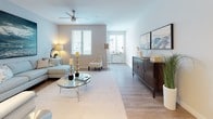 This single-floor residence showcases a thoughtfully designed open-concept layout finished with neutral tones and modern details throughout. Light wood-style flooring runs through the main living areas, creating a cohesive and bright aesthetic that extends from the entryway into the kitchen and living spaces.

The living area is spacious and well-lit, benefiting from multiple windows dressed with white horizontal blinds that allow generous natural light. A ceiling fan adds both function and style to the room, which comfortably accommodates a seating arrangement centered around a glass-topped coffee table and complemented by a substantial dark wood sideboard.

The kitchen is outfitted with sleek, high-gloss white cabinetry extending to the ceiling for ample storage, granite countertops, a subway tile backsplash, and a full suite of stainless steel appliances including a gas range, built-in microwave, and a French door refrigerator with an in-door ice and water dispenser.

The bedroom features plush carpeting, a ceiling fan, and a bright window, offering a calm and restful environment. The bathroom includes a white vanity with granite countertops, an undermount sink, and clean tile flooring. A dedicated in-unit laundry space houses a stacked washer and dryer for added convenience.