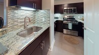 This single-floor residence offers a thoughtfully designed layout with a refined mix of materials and finishes throughout. The open living area makes a strong first impression with rich, dark hardwood flooring, crown molding, and track lighting that highlights the generous proportions of the space. Structural columns add an architectural detail while defining the flow between living areas.

The kitchen is a standout feature, outfitted with dark espresso cabinetry, granite countertops, a mosaic glass tile backsplash, stainless steel appliances including a gas range and built-in microwave, and a deep undermount sink. A breakfast bar connects the kitchen to the main living space, where an in-unit washer and dryer are also conveniently accessible.

The bedroom is bright and comfortable, with plush wall-to-wall carpeting, neutral walls, and a sliding glass door that opens to an outdoor view of surrounding greenery, allowing ample natural light to fill the room. A spacious walk-in closet with built-in wire shelving and hanging rods provides excellent storage.

The bathroom continues the home's consistent aesthetic with large-format tile flooring, a dark wood vanity with granite countertop, a framed mirror, and Hollywood-style vanity lighting. Clean lines and neutral tones create a calm, polished atmosphere throughout the entire residence.